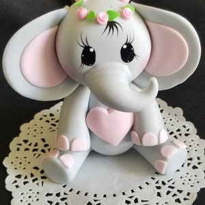 May include: A gray elephant figurine with pink ears, a pink heart, and a pink flower crown. The elephant is sitting on a white doily.