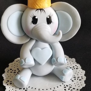 May include: A light blue and gray elephant cake topper with a yellow crown, holding a light blue heart. The elephant has big eyes and a small, curled trunk.