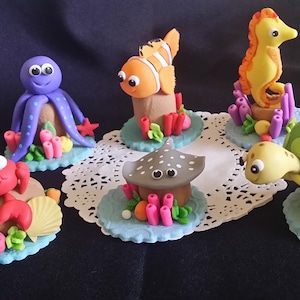 以下が含まれることがあります： カニ、タコ、タツノオトシゴ、カメ、エイ、クマノミなど、カラフルなフォンダン製の海の生き物のセットです。各生き物は、サンゴと海藻のアクセントが付いた小さな丸い台座の上に置かれています。