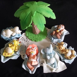 May include: A collection of baby animal figurines, including an elephant, giraffe, lion, monkey, and elephant, each resting on a blue blanket. A green palm tree is in the center. The figurines are made of a light-colored material.