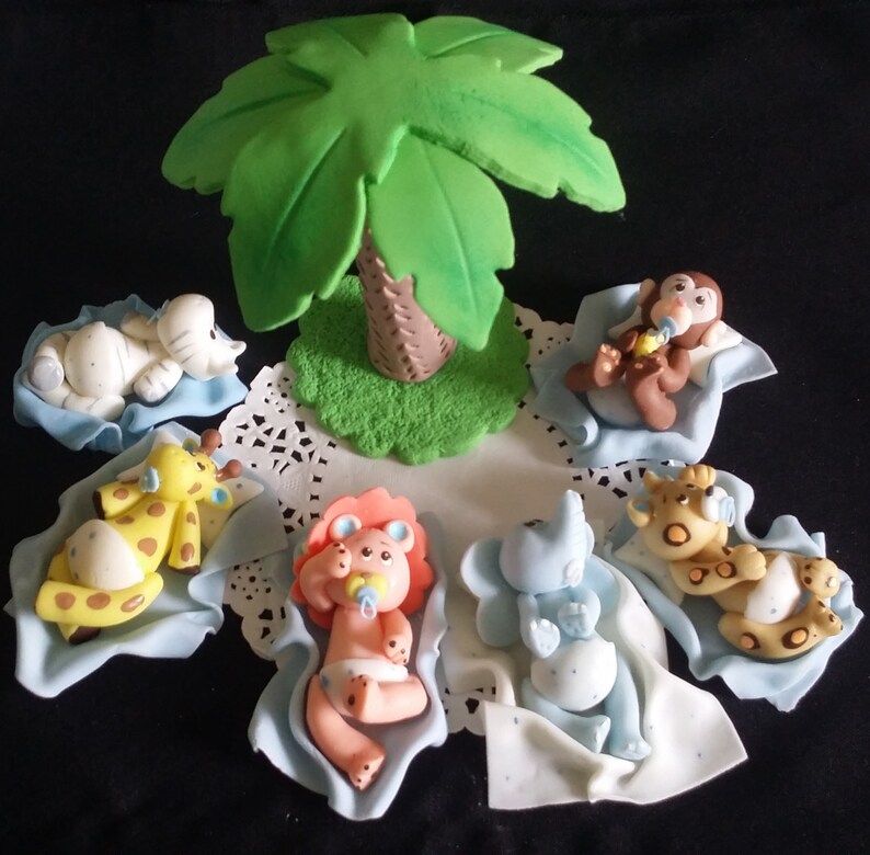 May include: A collection of baby animal figurines, including an elephant, giraffe, lion, monkey, and elephant, each resting on a blue blanket. A green palm tree is in the center. The figurines are made of a light-colored material.