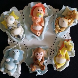 May include: A collection of six baby animal figurines, including a zebra, lion, cheetah, elephant, monkey, and giraffe. Each figurine is lying on a light blue blanket with a pillow. The figurines are made of a smooth material and are brightly colored.