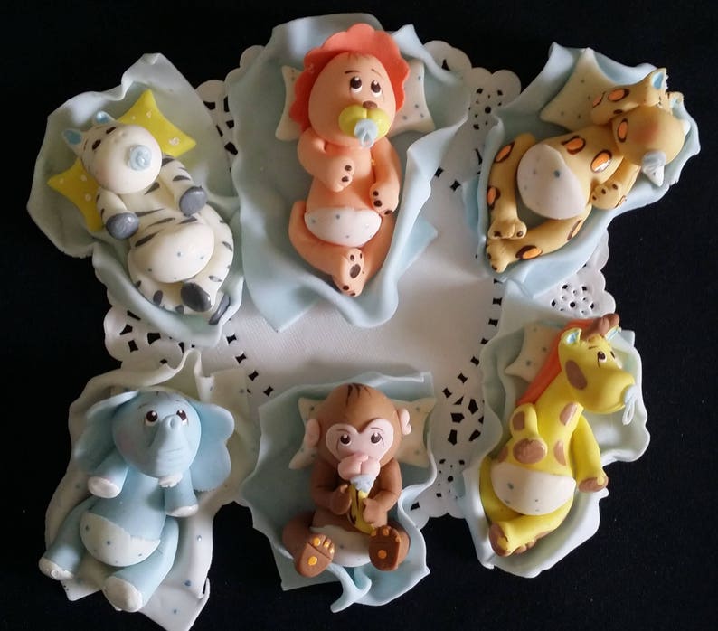 May include: A collection of six baby animal figurines, including a zebra, lion, cheetah, elephant, monkey, and giraffe. Each figurine is lying on a light blue blanket with a pillow. The figurines are made of a smooth material and are brightly colored.