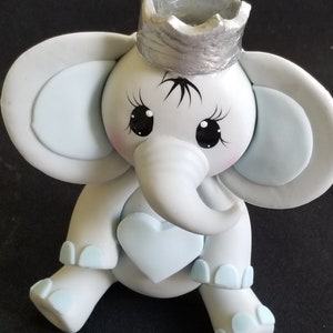 May include: A light blue and white elephant figurine wearing a silver crown. The elephant has a heart-shaped design on its chest and blue accents on its feet.