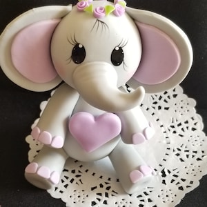 May include: A gray elephant figurine with pink ears and a purple heart. The elephant is wearing a flower crown with green leaves and purple flowers.
