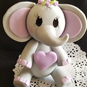 May include: A gray fondant elephant cake topper with pink ears, a purple heart, and a flower crown. The elephant has painted eyes and eyelashes.
