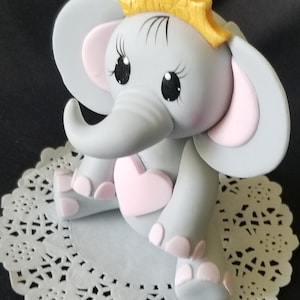 May include: Gray elephant cake topper with pink ears and a pink heart. The elephant is wearing a gold crown.