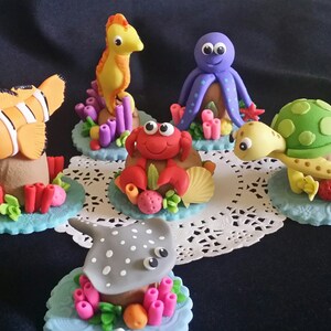 以下が含まれることがあります： ヒトデ、タコ、カニ、ウミガメ、クマノミ、エイなど、カラフルなフォンダンでできた海の生き物のセットです。それぞれの生き物は、サンゴや海藻の飾り付けが施された小さなケーキベースの上に置かれています。