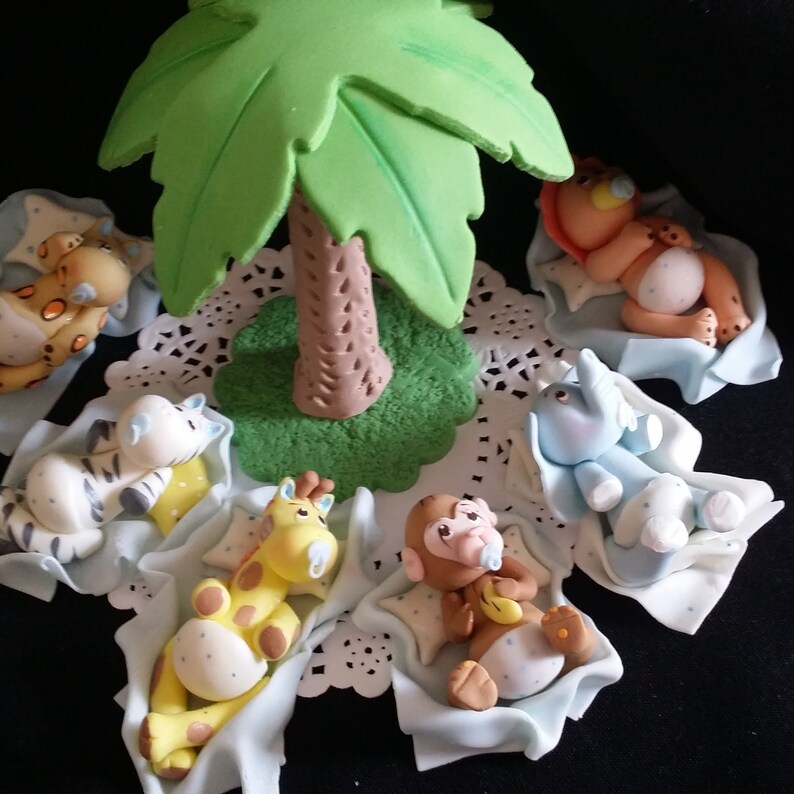 May include: A collection of fondant animal figurines, including a giraffe, zebra, lion, elephant, and monkey, each resting on a light blue blanket. A green palm tree is in the center. The figurines are on a white doily.