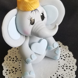 May include: A gray elephant cake topper with a blue heart and a gold crown. The elephant has blue ears and blue feet.