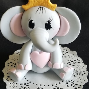 May include: A gray elephant cake topper with pink ears and a pink heart. The elephant is wearing a gold crown and has black eyes with eyelashes.