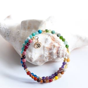Puede incluir: Un brazalete de cuentas multicolores con un dije de Om dorado. El brazalete está hecho con varias piedras preciosas, incluyendo cuentas rojas, naranjas, amarillas, verdes, azules y moradas. El brazalete está descansando sobre una concha blanca.