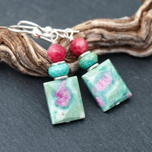 Op de afbeelding: Een paar oorbellen met rechthoekige groene en roze stenen hangers. Elke oorbel bevat een ronde rode kraal en een ronde turquoise kraal, verbonden door zilverkleurige metalen onderdelen en zilverkleurige oorhaken.