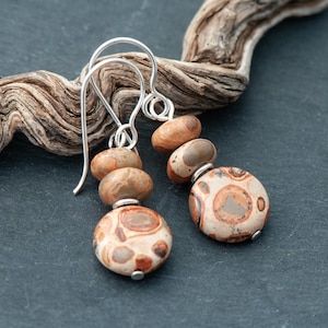 Peut inclure: Boucles d'oreilles pendantes composées de perles en pierre naturelle dans des tons marron, beige et crème. Les boucles d'oreilles sont dotées de crochets argentés et sont présentées sur un fond sombre avec un morceau de bois flotté.
