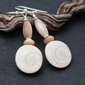 May include: A pair of dangle earrings featuring a spiral design. The earrings have a light beige color with silver-tone hooks. The earrings are composed of a light beige oval-shaped spiral design, with a light beige elongated bead and a small round bead.