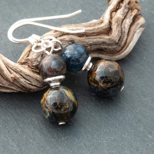 Op de afbeelding: Close-up van twee paar oorbellen met zilveren haken. Elke oorbel heeft twee bolvormige kralen in tinten blauw, bruin en goud, gescheiden door een zilveren accent. De oorbellen rusten op een stuk drijfhout.