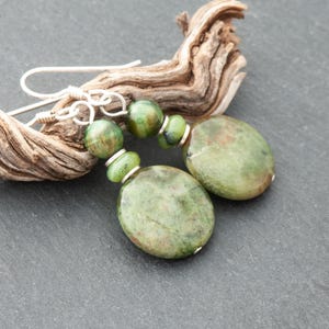 May include: Handmade green stone earrings with silver hooks. The earrings feature large, oval-shaped stones and smaller, round beads in shades of green. The earrings are displayed on driftwood against a dark grey background. The earrings are a unique piece of jewellery.