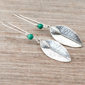 Peut inclure: Une paire de boucles d'oreilles en forme de feuilles argentées avec un motif texturé. Chaque boucle d'oreille comporte une perle verte et un long crochet fin. Les boucles d'oreilles sont présentées sur une surface en bois.