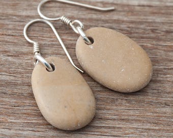 May include: A pair of handcrafted earrings featuring smooth, oval-shaped beige stones. The stones are suspended from silver-colored ear wires. The earrings are displayed on a wooden surface, showcasing their natural aesthetic.