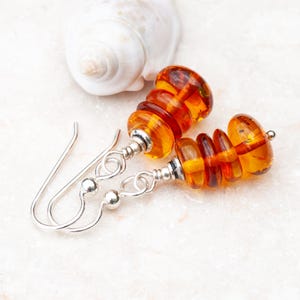 May include: A pair of amber-colored earrings with a silver hook closure. The earrings feature a stacked design of graduated amber beads, with silver accents between each section. A white seashell rests in the background.