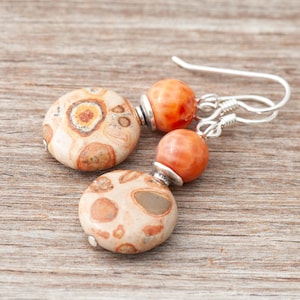 Puede incluir: Un par de pendientes hechos a mano con cuentas de piedra natural. Los pendientes tienen una combinación de cuentas redondas beige y marrón moteadas, y cuentas esféricas naranjas. Los pendientes cuelgan de ganchos plateados.
