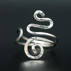 Puede incluir: Un anillo de plata con un acabado martillado y un diseño abstracto único. El anillo está hecho de dos bandas entrelazadas, una de las cuales forma una espiral.