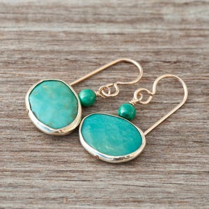 Peut inclure: Une paire de boucles d'oreilles turquoise et argent. Chaque boucle d'oreille présente une grosse pierre turquoise ovale sertie dans un cadre argenté, avec une petite perle verte et un crochet en fil doré. Les boucles sont posées sur une surface en bois.