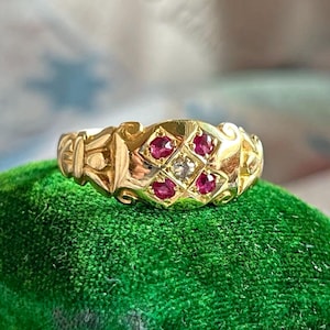 Puede incluir: Un anillo de oro con un diseño de diamantes y piedras preciosas de rubí. El anillo presenta un diamante central rodeado de cuatro rubíes en un patrón en forma de diamante. La banda tiene un diseño ornamentado y texturizado.