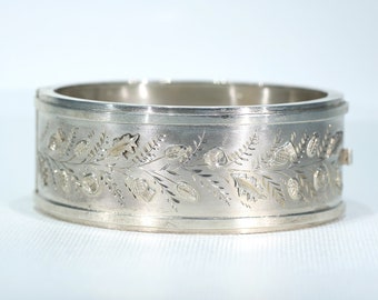 Puede incluir: Un brazalete de plata con un diseño floral grabado. El diseño presenta hojas y flores.
