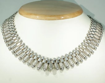 May include: A silver necklace with a geometric design. The necklace is made of small, cylindrical beads that are connected together in a repeating pattern. The beads are textured and have a slightly hammered finish.