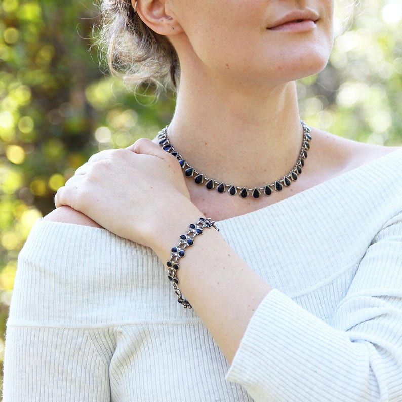 Pu&ograve; includere: Un set di collana e braccialetto in argento con pietre nere a forma di lacrima. La collana &egrave; di stile choker e il braccialetto &egrave; una catena delicata.