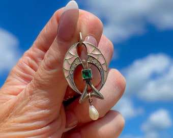 May include: A silver pendant with a pink and white enamel design, featuring a green gemstone and a pearl drop. The pendant is in the shape of two crescent moons.