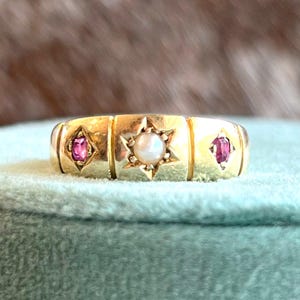 May include: A gold ring with a central pearl set in a starburst design, flanked by two pink gemstones in diamond-shaped settings. The ring has a rectangular band with a polished finish, showcasing a vintage aesthetic.