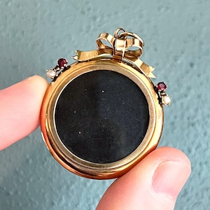 May include: A round gold pendant featuring a central black circular inlay. The top of the pendant is adorned with a gold bow motif. On each side, there is a small cluster consisting of one red gemstone and one white pearl set in gold metal.