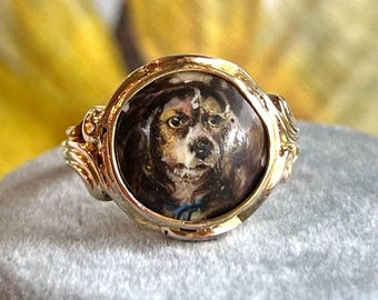 May include: A gold-toned ring featuring a detailed portrait of a dog within a circular setting. The dog's fur is depicted in shades of brown, black, and white, with a blue collar. The ring has ornate, swirling details on the band.