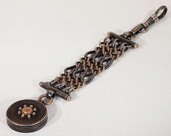 Op de afbeelding: Een zakhorlogeketting van zilver en koper met een ronde hanger. De ketting heeft een geweven ontwerp en is bevestigd aan een draaibare sluiting.