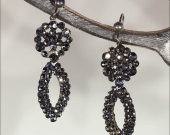 May include: A pair of silver earrings with a teardrop-shaped design. Each earring features a cluster of black rhinestones in the shape of a flower and a teardrop.