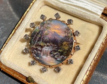 May include: A gold and silver brooch with a painted landscape scene of a waterfall in a forest. The brooch is surrounded by small diamonds.