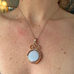 Pode incluir: Um colar com pingente dourado com um centro circular azul claro. O pingente tem um design ornamental e rodopiante acima do elemento circular. A corrente é delicada e dourada.