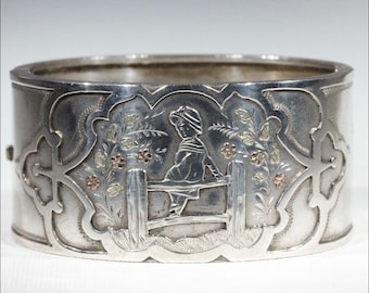 Puede incluir: Un brazalete de plata con un diseño grabado de un niño sentado en una valla con flores. El brazalete está decorado con detalles dorados.