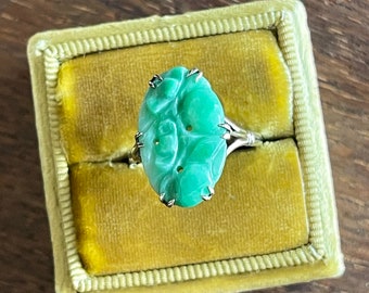 May include: A green jade ring with a carved floral design set in a gold band. The ring is displayed in a yellow velvet ring box.