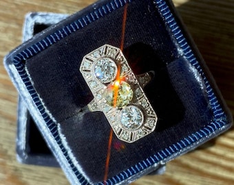 May include: A silver ring with two large diamonds and a smaller yellow diamond in the center. The ring is in a blue velvet ring box.