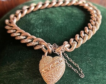 May include: A gold-toned bracelet with a heart-shaped lock. The bracelet features a thick chain design. The heart-shaped lock is intricately detailed with patterns and a diagonal stripe. A small chain hangs from the lock.