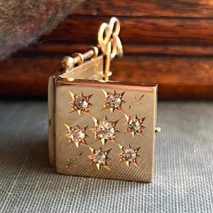 May include: A gold-colored, rectangular locket with a hinged opening. The front of the locket features nine starburst designs, each with a central, clear gemstone. The locket has a small key-shaped clasp and a textured surface.