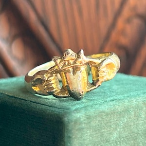 May include: A gold-colored ring with an ornate design. The ring features a central shield-like element flanked by detailed, sculpted elements. The ring's band is smooth and rounded, and the overall design suggests a vintage or antique style.