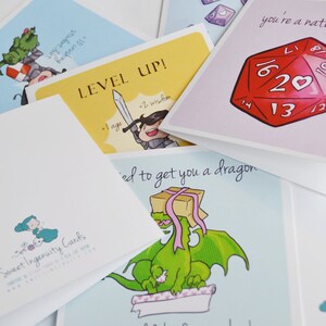 Puede incluir: Un conjunto de tarjetas de felicitaci&oacute;n con varias ilustraciones y texto. Una tarjeta presenta un drag&oacute;n verde con una cinta rosa, otra tarjeta tiene un caballero con una espada y una tercera tarjeta tiene un dado rojo de 20 caras.