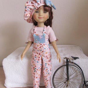 May include: A doll with brown hair wearing a pink floral bonnet with a blue bow, a light pink t-shirt, and matching pink floral overalls with blue accents. The doll wears floral sneakers and stands next to a miniature black penny-farthing bicycle.