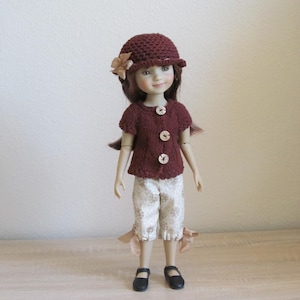 May include: A doll wearing a brown knitted cardigan with buttons, white patterned shorts, and a brown knitted hat with a flower accent. The doll has brown hair and blue eyes.