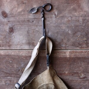 May include: A brown leather apron with a large oval patch hangs on a black metal hook shaped like a leaf. The hook is attached to a wooden wall.