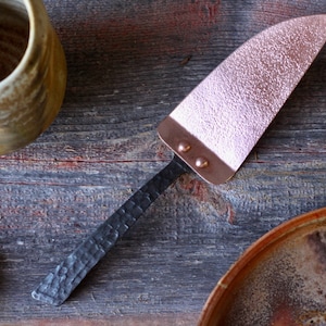 May include: A copper cake server with a hammered black metal handle. The server is on a rustic wooden surface.